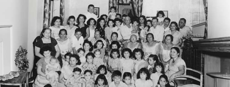 A black and white family photo with over 30 people posing for a group shot. The ages range from young children to older adults. The photo is captured in a living room setting. There are windows and curtains in the background. Some children are sitting crossed-legged or kneeling in the front, other family members are sitting on chairs in the middle, and many are standing in the back. 