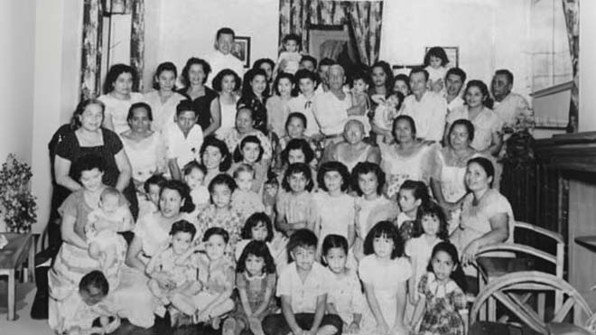A black and white family photo with over 30 people posing for a group shot. The ages range from young children to older adults. The photo is captured in a living room setting. There are windows and curtains in the background. Some children are sitting crossed-legged or kneeling in the front, other family members are sitting on chairs in the middle, and many are standing in the back. 