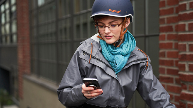 This is an image of a woman on a bike outside. She's in front of a brick building. She's wearing a gray rain jacket, a blue scarf and a helmet. She's looking down at her phone.