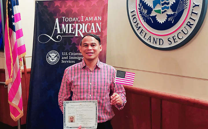 BECU employee Jom G smiles as he holds up a certificate as he receives U.S. citizenship. In the background is an American flag, the Homeland Security logo and a citizenship backdrop. He is holding up a small American flag.