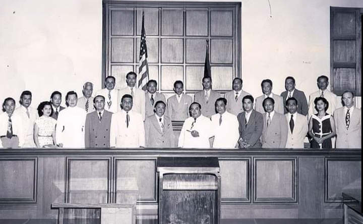 Black and white capture of the first Guam legislature. There are 25 people standing for the group photo. They are in a courtroom setting. In the background are two flags and wooden windows. There is a podium in the center.