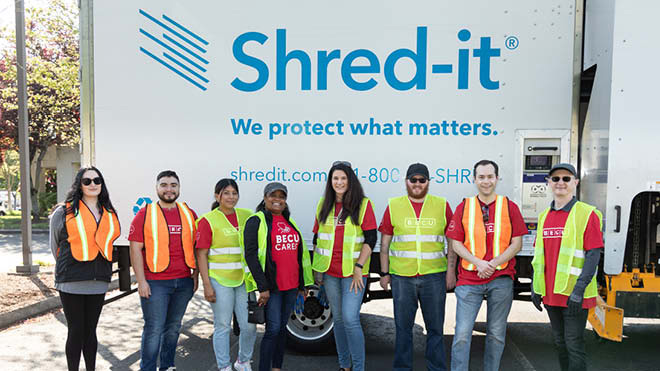 A group of smiling people wearing work vests in front of a truck with the Shred-it® logo.