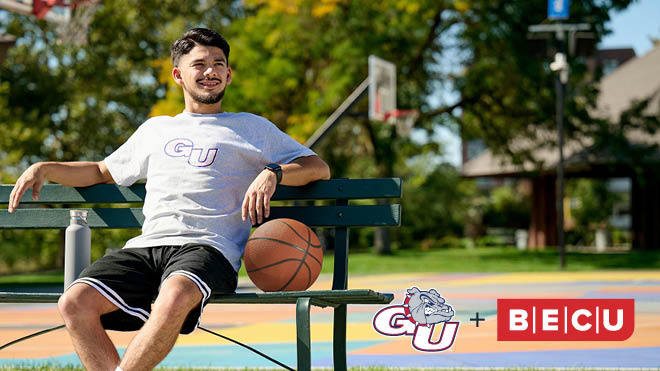 A smiling man wearing a Gonzaga University shirt sitting on a bench with a basketball. Gonzaga University plus BECU logos.