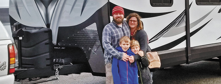 A father, mother and their two boys pose for a family photo in front of their RV, which is hitched to their pickup truck.