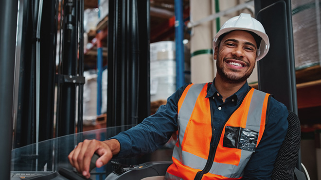 Photo of a smiling man driving a forklift and wearing personal protective equipment.