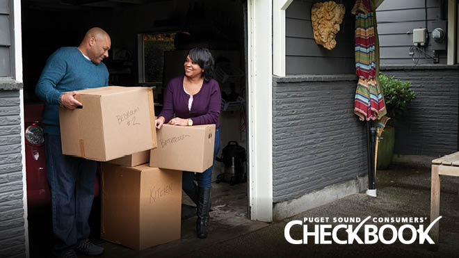 A man and woman stand in their garage doorway. The man is carrying a cardboard box labeled "BEDROOM # 2" in black marker. A woman, who is smiling at him, rests her hands on top of a stack of labeled cardboard boxes. A white text logo in the lower right says "Puget Sound Consumers' Checkbook."