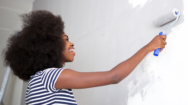 A woman smiles as she paints a white wall gray using a roller brush.