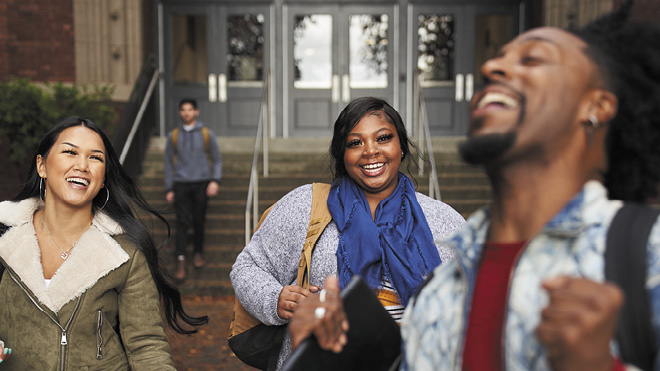 Three college students laugh as they walk away from a building on campus together.