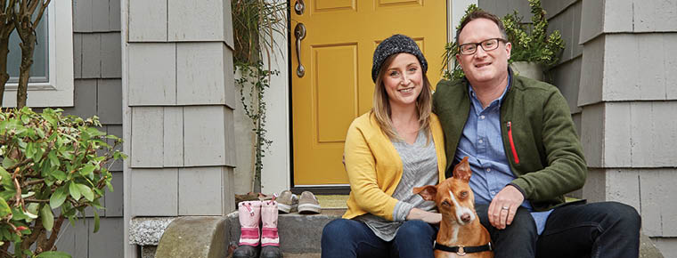 A woman, man and dog sit on the front stairs outside their gray house. The bright yellow front door is ajar behind them.
