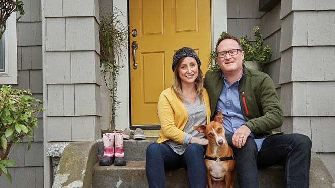 A woman, man and dog sit on the front stairs outside their gray house. The bright yellow front door is ajar behind them.
