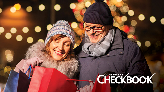 Couple standing shoulder to shoulder together outside, dressed in warm winter clothes, smiling and looking down inside of a red shopping bag, with holiday lights visible in the background. "Consumers' Checkbook" logo in the bottom right corner.