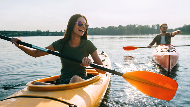 Two people kayaking in orange boats with orange paddles on a sunny day. A woman in the foreground is smiling, looking in the direction that the man in the boat behind her is pointing. The sun is shining on the calm water.