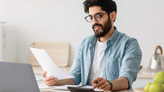 A man sits in his kitchen looking at papers he's holding in his right hand and punching buttons on a calculator with his left hand. A laptop is open on the table in front of him.