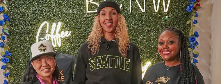 Three women standing together in front of a green, leaf-covered wall with a neon sign that says "Coffee" on it. The woman on the left is Intentionalist CEO and founder Laura Clise, wearing a Seattle Sonics hat. The woman in the middle is Seattle Storm Center Mercedes Russell, wearing a sweatshirt that says "Seattle." The woman on the right is Black Coffee Northwest co-owner DarNesha Weary, wearing a "Black Coffee Northwest" t-shirt.