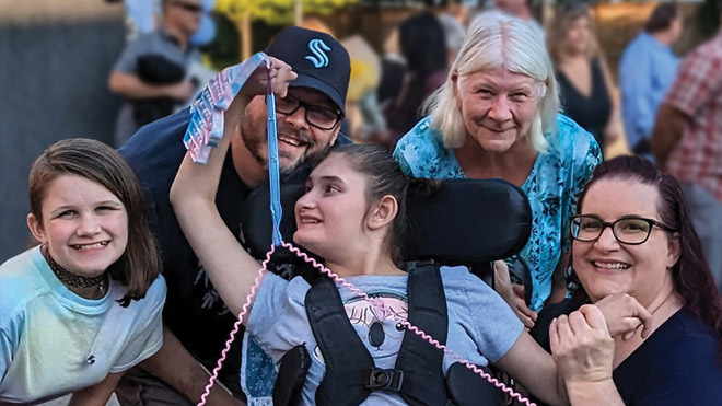 Young girl in a wheelchair smiles as she looks back at four other members of her family, who are surrounding the young girl on all sides while posing and smiling for the photo.