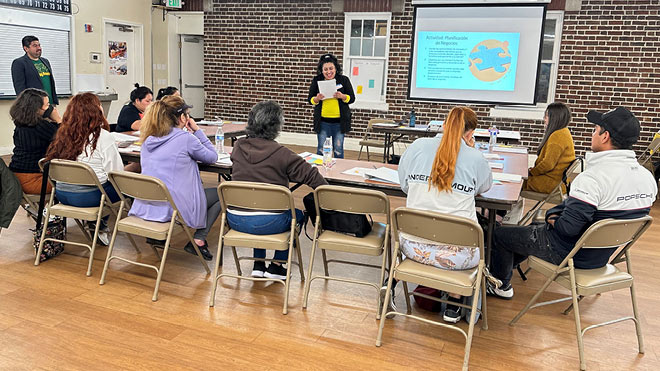 A group of nine individuals are seated inside at a table, while one person is standing and presenting in front of the group. The person presenting is holding a piece of paper and reading. To the left of the group another individual is standing and listening. The background displays a projector screen and a brick wall.