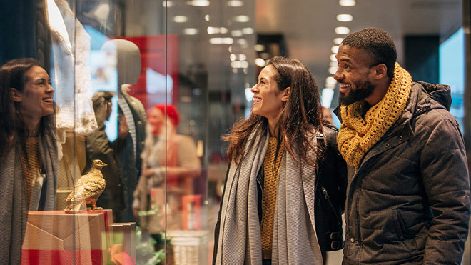 A couple are shopping and smiling as they look into a window together. The two are wearing winter clothes. Both are wearing scarves and are shopping indoors in a mall-like setting. 
