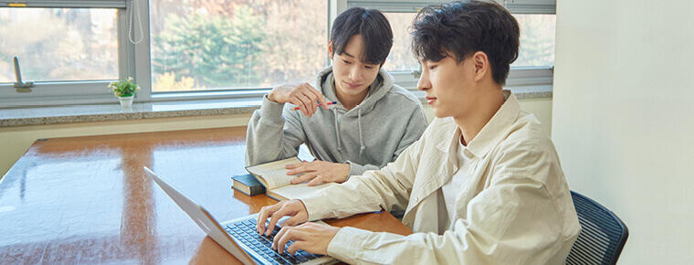 Two young college-aged students sitting at a table inside located in front of large windows. Trees are in the background outside. The two students are looking together at the screen of a laptop that's in front of one of the students, who is typing on the laptop keyboard. The other student not in front of the laptop is holding a pen in one hand and has a book in their other hand resting on the table, open to a page.
