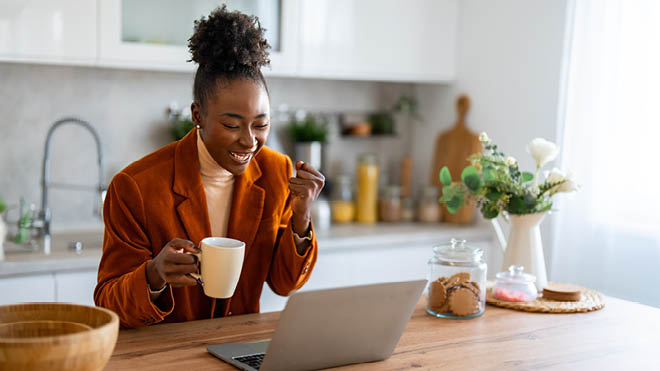 A woman looks down at her laptop and is smiling. One of her hands is raised happily in a clench position suggesting an achievement concept. In her other hand is a cup of coffee. She's seated in a kitchen setting. On the table are flowers in a vase. A sink, cutting board, and various kitchen appliances are in the background. She is wearing an orange blazer.