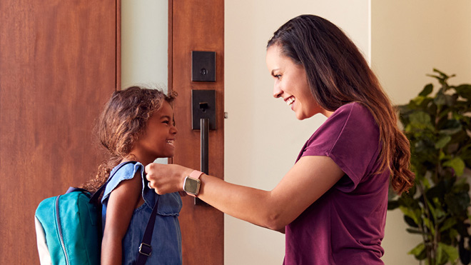 Young girl sporting a light blue backpack smiles as she stands in front of her smiling mother, who has placed her left hand on the young girl's shoulder, a showing of support as the young girl readies to leave through the front door of their home to go back to school.
