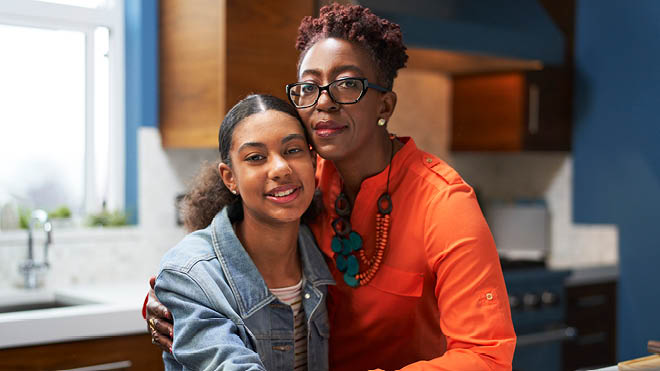 A mother and her teen daughter stand together in their kitchen facing the camera for a photo. The mom has her arm around the teen and is resting her cheek against her daughter's head.