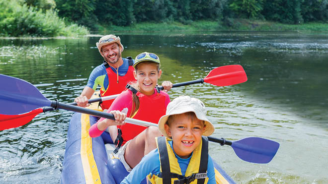 Three people on a kayak in a lake. A man wearing a red life vest in the back, a boy wearing a red life vest in the middle and a boy wearing a yellow life vest in the front. They are holding paddles and smiling at the camera.