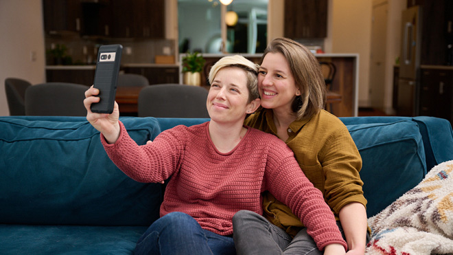 Two women sit close together on couch, holding hands. Woman on the left is holding phone up to take a selfie.