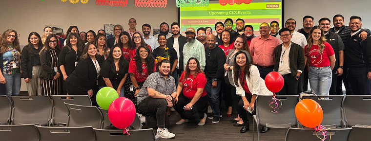 A large group of BECU's Latinx Employee Resource Group (ERG) are gathered together inside at an employee event. The group is standing in front of a screen posing for a photo. There are balloons and decorations in the background.