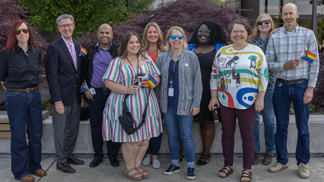 10 BECU employees pose for the camera, smiling and standing side by side outside of Tukwila Financial Center for a recent Pride flag raising. Some employees pictured are holding and waving small Pride flags.