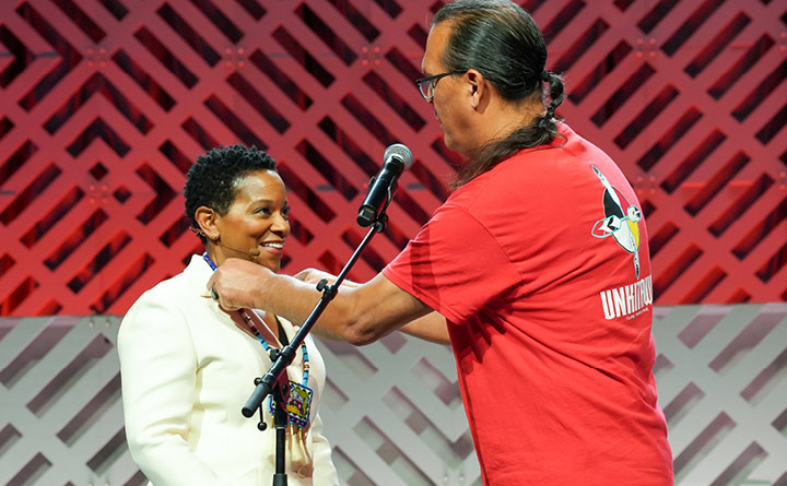 Kyle Schierbeck, project executive for Unkitawa, and Beverly Anderson, BECU CEO, are standing on a stage in front of microphone. Kyle is putting a necklace around Bev's neck. Bev is smiling.