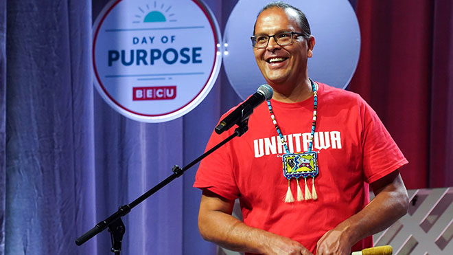 Kyle Schierbeck, project executive for Unkitawa, is standing on a stage in front of microphone smiling. He is wearing a red shirt that says Unkitawa and is wearing a large beaded colorful necklace. Behind him is a BECU sign that says "Day of Purpose BECU."