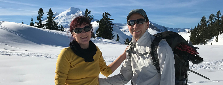 A couple pose in the snowy mountains for a photo. The two are standing and smiling and both people are wearing sunglasses.  There are mountains and evergreen trees in the background. The sky is blue. The woman is wearing a yellow long-sleeved shirt with a scarf, and the man is wearing a long-sleeved white shirt and has a hat and backpack on. 