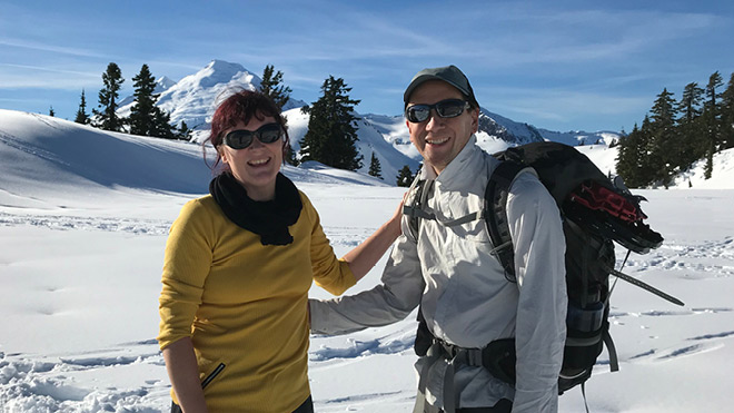 A couple pose in the snowy mountains for a photo. The two are standing and smiling and both people are wearing sunglasses.  There are mountains and evergreen trees in the background. The sky is blue. The woman is wearing a yellow long-sleeved shirt with a scarf, and the man is wearing a long-sleeved white shirt and has a hat and backpack on. 