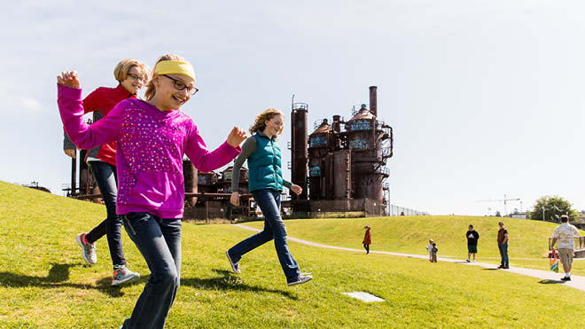 This is an image of three younger girls running at Gas Works Park in Seattle, Washington. They are all smiling and the girl in the front has her hands in the air. She is wearing a pink sweatshirt and a yellow headband. The other two girls are wearing red and blue sweatshirts. In the background are people and children hanging out at the park. Also in the background is the gasification plant. The grass is bright green and the sky is light blue.