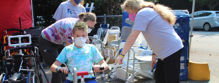 A young boy is sitting on an adaptive bike, while three individuals help to make sure the bike fits correctly. All three women are looking at the bike. Two are wearing gray shirts and one is wearing a tie dye shirt. In the background is other assistive devices such as wheelchairs, walkers and other equipment. They are outside under a red canopy tent. Behind the group are trees and a couple parked cars.