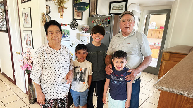 A family stands together in their kitchen, smiling for a photo. There is a grandfather, grandmother and three grandchildren. There are several framed photos, a refrigerator and vase with flowers on the floor behind them. 