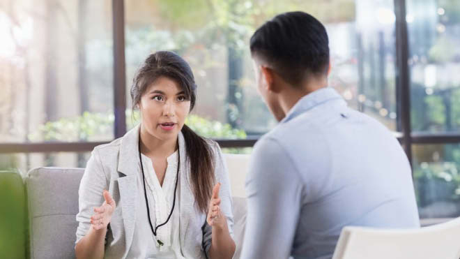 A woman is seated on a couch as she counsels a person who is also seated in a therapy looking setting. She has both her hands raised a bit lower than shoulder length as if she's explaining something. She is wearing a black lanyard, a gray blazer and has long brownish black hair with a side pony tail. The man, who is facing away from the camera has black hair and is wearing a light blue button up. In the background are large windows showing bright light and trees.