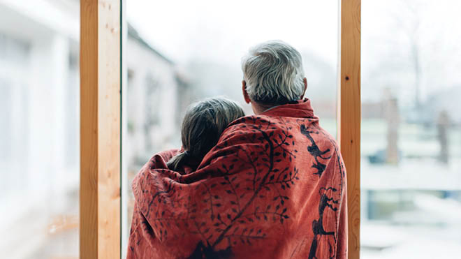 An elderly couple looks out of a window, holding each other with a red blanked draped over their shoulders. 