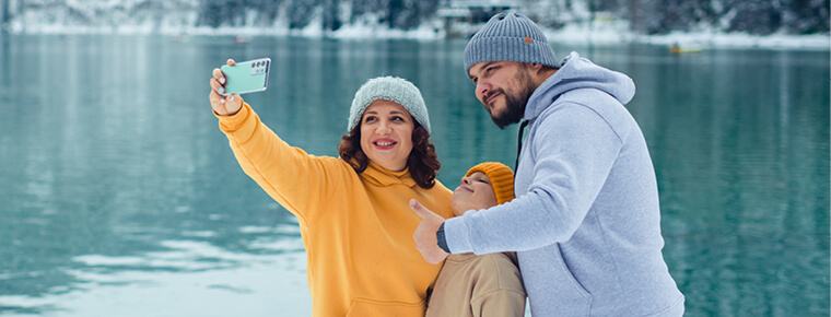 A family is smiling and posing for a selfie. They are outdoors by a lake. The lake and background are set in the wintertime and there is light snow in the background. A woman is holding up her phone to take the selfie. She has a blue beanie and yellow sweatshirt on. The man has a gray beanie and a gray sweatshirt on and is holding a thumbs up. The son is standing in between the couple and has a yellow beanie and is wearing a tan sweatshirt. 