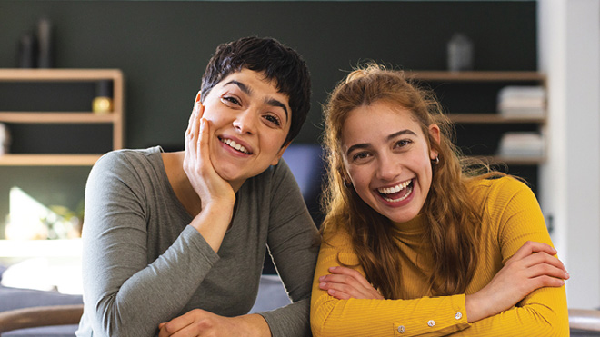 Two women are smiling inside a living room setting facing the camera while seated and posing for a photo. The woman on the left has dark, short hair. Her hand is resting on her chin as she smiles. She is wearing a gray long sleeve shirt. The woman on the right has long red hair. She is wearing a yellow long sleeve shirt. Her arms are crossed as she smiles big.  The background is blurred out, but shows some shelving in a living room setting.