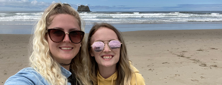 A mother and daughter standing on a sandy beach, both smiling and wearing sunglasses. The ocean, white waves, blue sky and a large rock are visible in the background. 