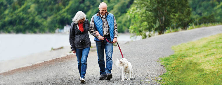 A couple is outdoors on a path walking their dog. The man is wearing a blue vest, a long sleeve plaid shirt and blue jeans. The woman is wearing a black coat with a red shirt underneath. She is also wearing blue jeans. The dog is white. In the background are trees and water. 