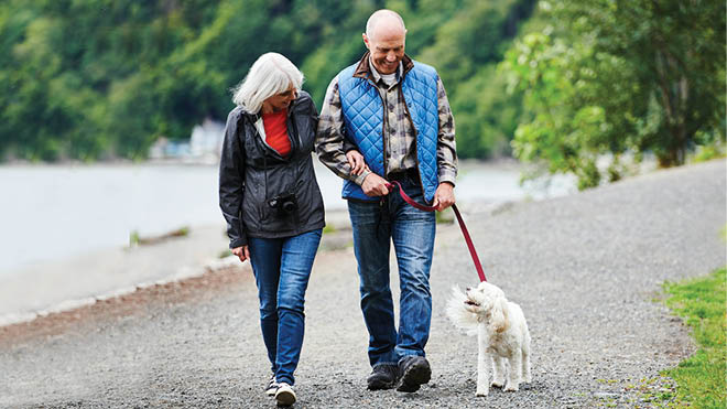A couple is outdoors on a path walking their dog. The man is wearing a blue vest, a long sleeve plaid shirt and blue jeans. The woman is wearing a black coat with a red shirt underneath. She is also wearing blue jeans. The dog is white. In the background are trees and water. 