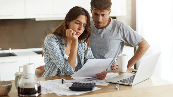 A couple is looking at paperwork in their kitchen. A woman is holding paperwork in her hand while her other hand is rested on her chin in a thinking posture. The man is behind her and is also looking at the paper with one of his hands on his hip. They are in a indoor kitchen setting. The woman appears to be sitting at a kitchen counter, while the man is standing. There are two mugs on the countertop and a computer is open as well. 