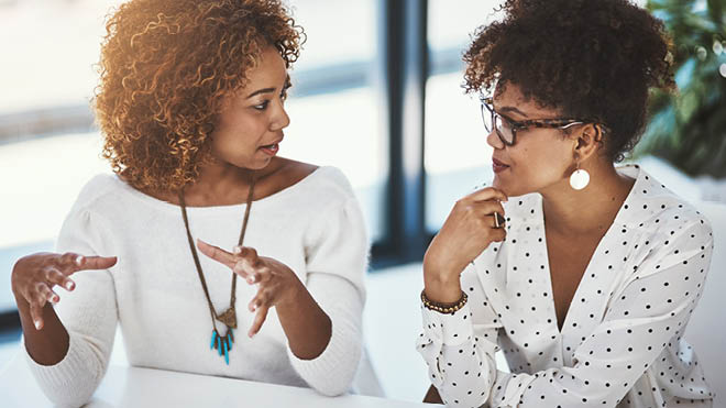 Two women are indoors, seated in an office setting. The woman on the left has brown curly hair. She's holding up both hands, in a descriptive position. She is wearing a white long sleeve shirt and a necklace. The woman on the right has curly dark brown hair. She's wearing her hair up. She has glasses and a white dangly earring. She's wearing a white blouse with black polka dots. She has one hand on her chin, like she's listening. In the background are windows and a plant.