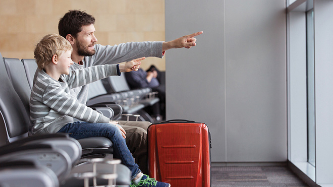 This is an image of a father and son in an airport. They are sitting down in chairs and looking out a window and pointing outside. In the photo in a red suitcase. The son is wearing a gray stripped shirt and has blonde hair. The father is wearing a gray shirt and has brown hair. 