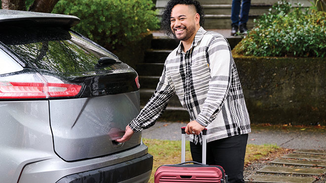 A smiling person starts to open the hatch back of a silver car parked in front of a building. He has a suitcase in his hand. He is wearing a black and white striped shirt. In the background is a sidewalk, green bushes and a staircase leading up to the front of a house. Another person is standing at the front of the house.