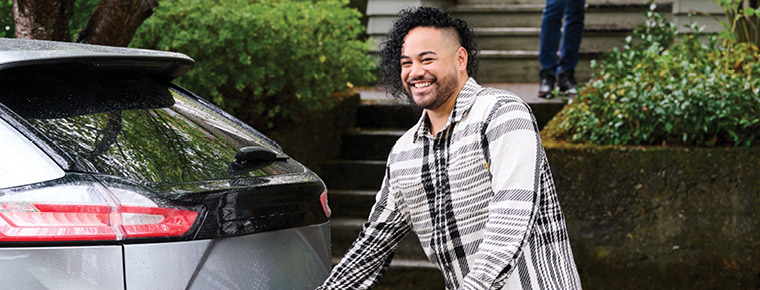 A smiling person starts to open the hatch back of a silver car parked in front of a building. He has a suitcase in his hand. He is wearing a black and white striped shirt. In the background is a sidewalk, green bushes and a staircase leading up to the front of a house. Another person is standing at the front of the house.