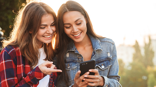 Two young women smile as they look at a phone together. The woman on the left is pointing at the phone as the other holds it. They are outdoors and there are trees in the background.
