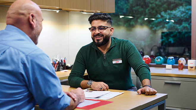 A BECU employee sits down with a member in an office at a neighborhood financial center. The employee is wearing a name tag and a green shirt. The member is facing away from the camera and he is wearing a blue shirt. In the background are desk cabinets, some office supplies like pens and pencils in containers at the employee's desk and a window facing trees.